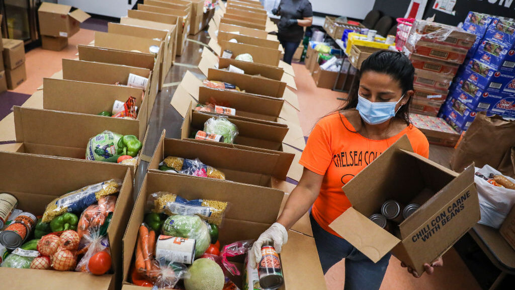 A woman wearing an orange shirt and blue surgical mask sorts canned foods into a long row of boxes ready for distribution at the Chelsea Collaborative, an activist organization in one of the cities in Massachusetts hardest-hit by Covid-19.