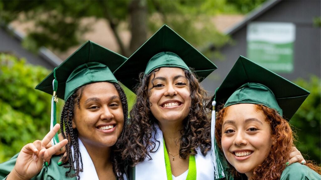 Three female high school graduates smile and embrace wearing green caps and gowns.