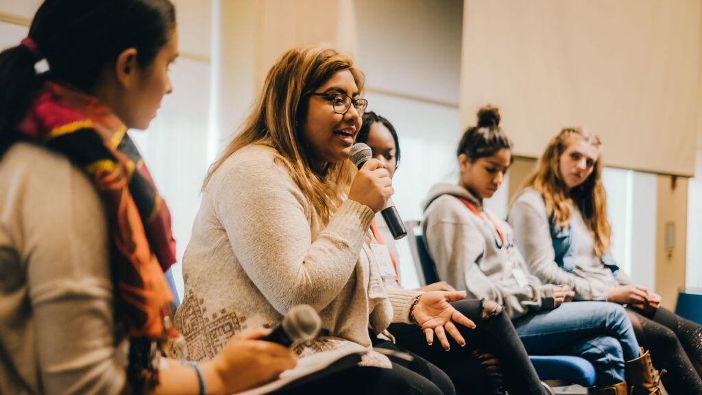 A group of high school students sit on a panel, one is holding a microphone and speaking.