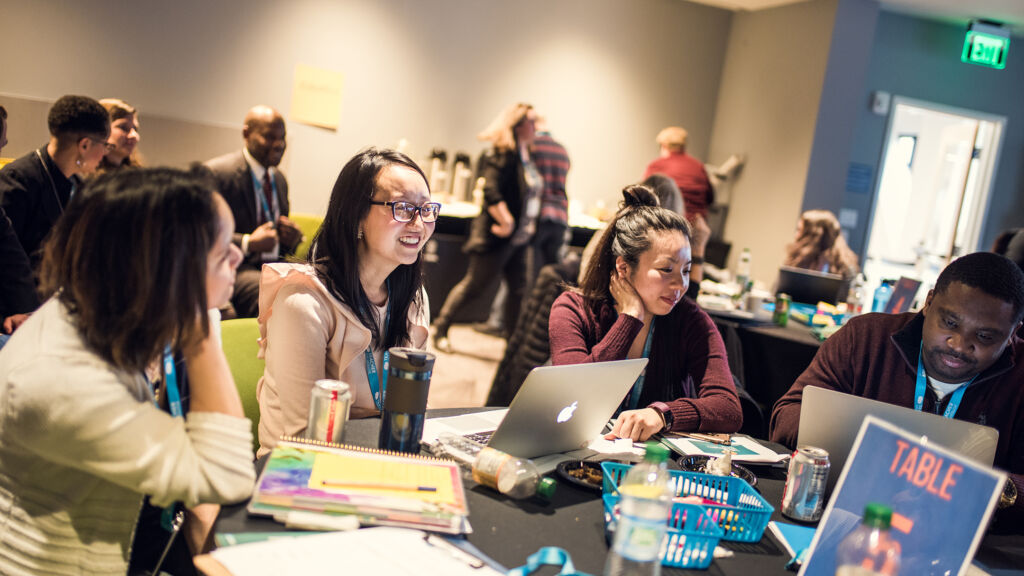 A group of teachers sitting at a table covered with papers and laptops. The teachers are talking and smiling.