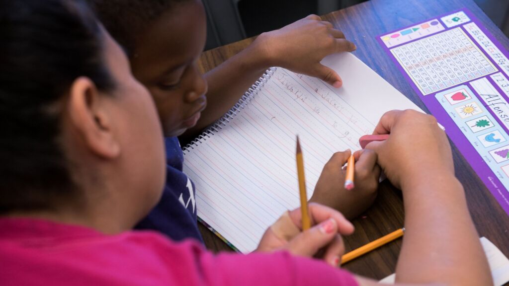 A teacher works with a young student on a handwriting assignment. The teacher leans in close with an eraser to help the student write a sentence about winter weather.