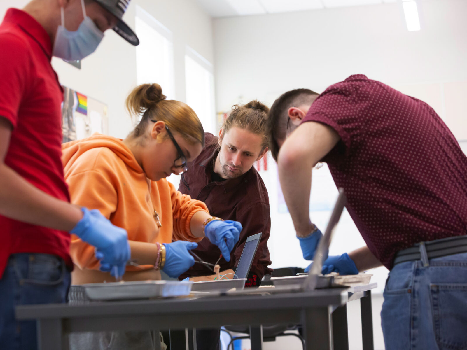 Three students wearing blue surgeon gloves are actively working in a high school classroom, dissecting chicken wings, while a teacher looks on supportively.