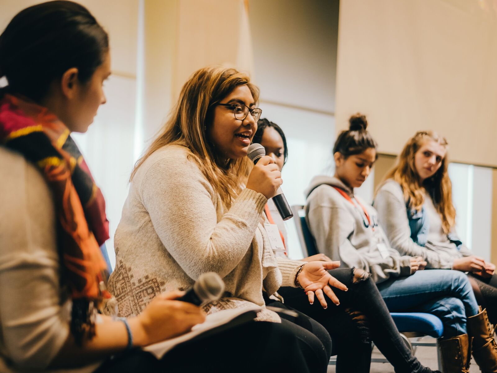 A group of high school students sit on a panel, one is holding a microphone and speaking.