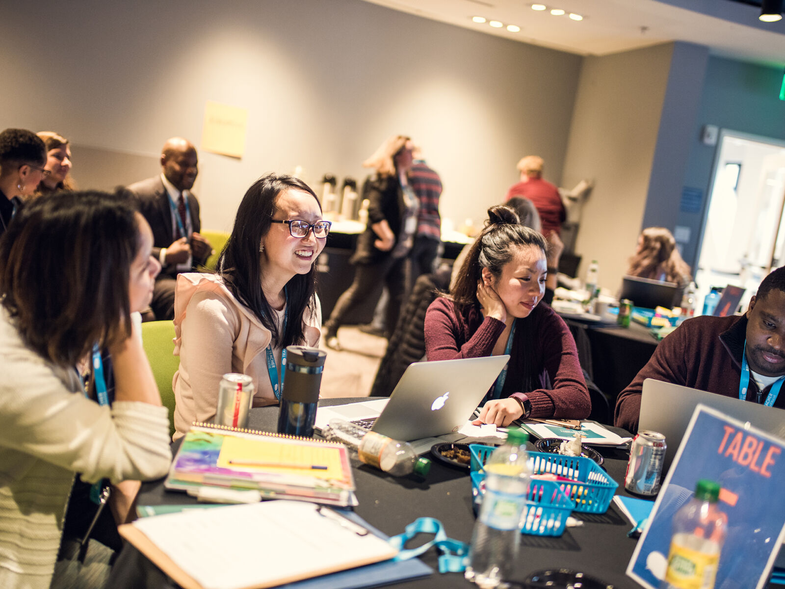 A group of teachers sitting at a table covered with papers and laptops. The teachers are talking and smiling.