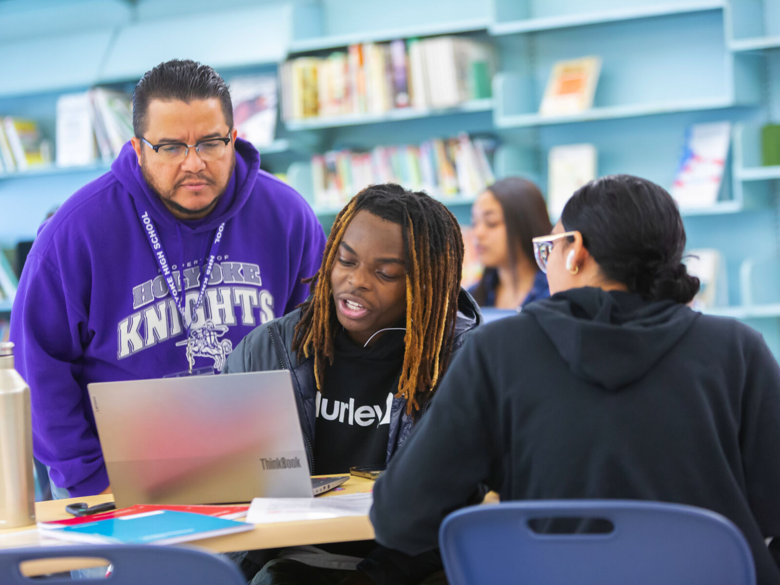 A teacher wearing glasses and a purple Holyoke sweatshirt looks over the shoulder of a student who is explaining something on his computer. They are in a school library with other students nearby.
