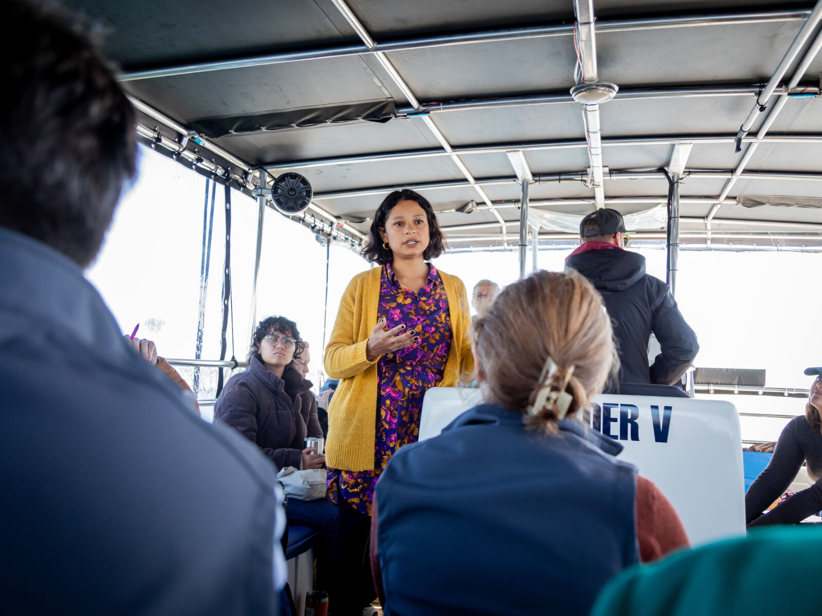 Woman wearing a gold sweater and patterned blouse speaks to a group of people sitting on a small boat while touring the Mystic River waterfront to discuss climate change.
