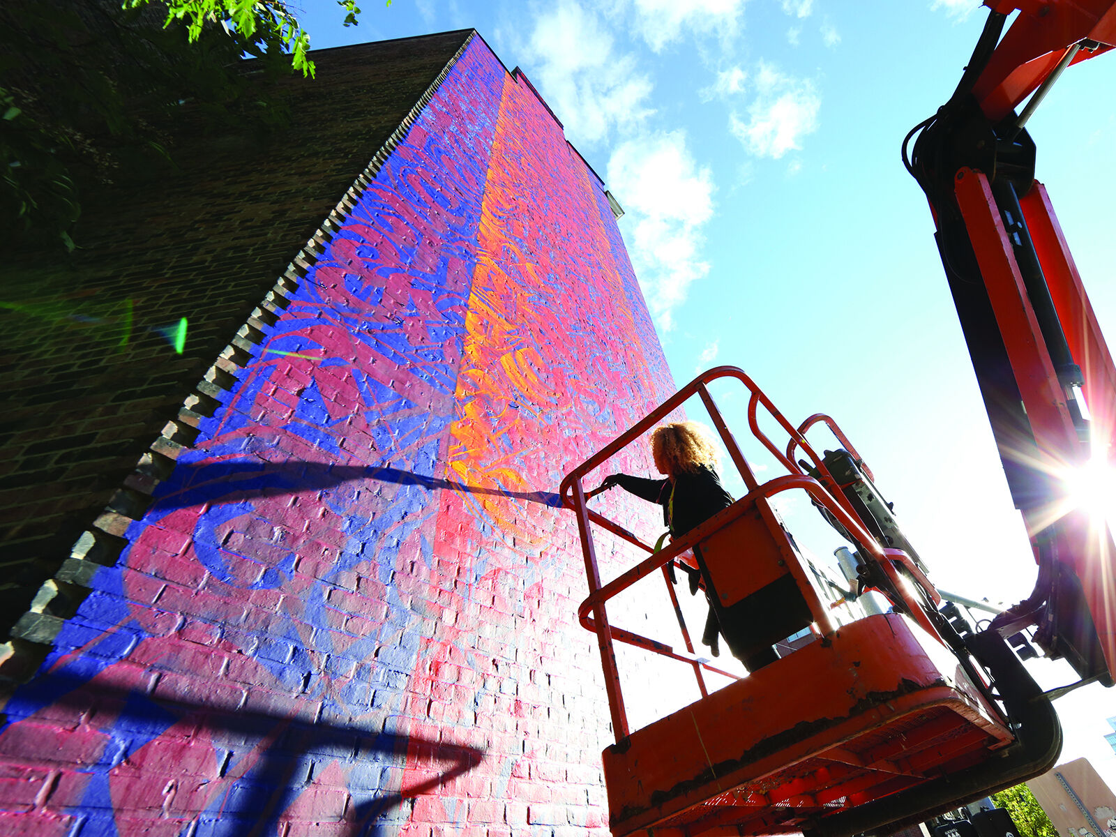 Looking up at a largescale purple and red mural while the artist stands on a lift and works on it.