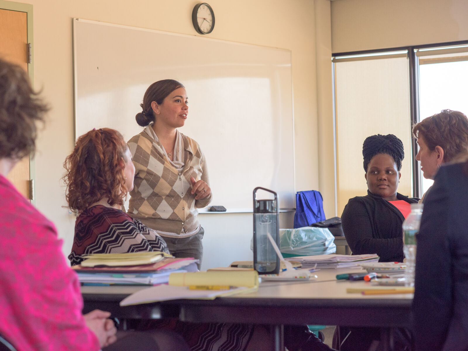 A Latina woman stands in front of a group of people who are sitting at desks and listening to her speak