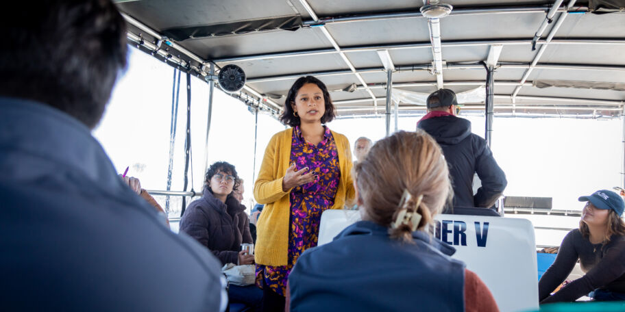Woman wearing a gold sweater and patterned blouse speaks to a group of people sitting on a small boat while touring the Mystic River waterfront to discuss climate change.