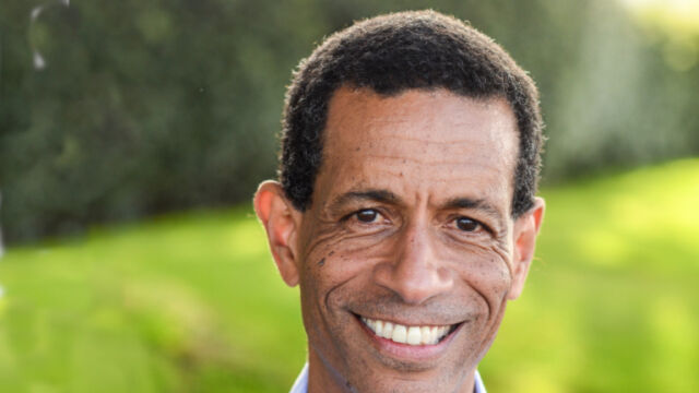 Mark Edwards wearing a gray suit jacket, blue button down shirt, smiling and standing in front of a brick wall