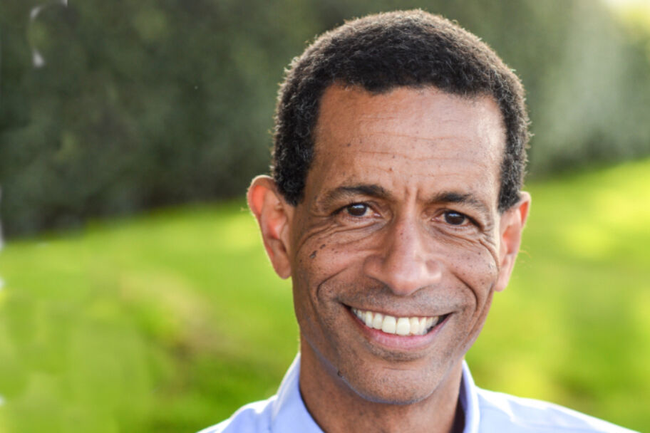 Mark Edwards wearing a gray suit jacket, blue button down shirt, smiling and standing in front of a brick wall